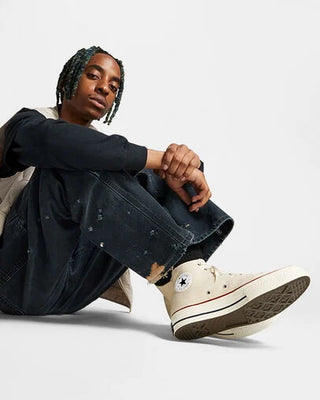 Person wearing white high-top sneakers with a brand logo, sitting on a light gray background