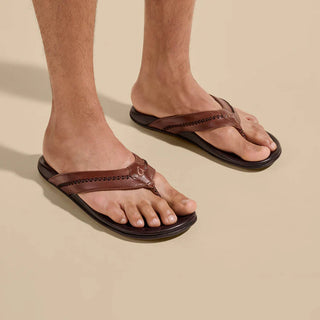 Olukai Mea Ola leather sandals in Tan and Dark Java with whipstitched straps and laser-etched footbed artwork.
