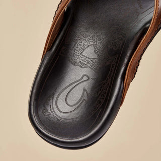 Olukai Mea Ola leather sandals in Tan and Dark Java with whipstitched straps and laser-etched footbed artwork.