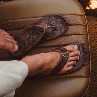 Side view of an OluKai Mekila sandal in Natural/Natural with brown leather straps, stitched edges, and decorative artwork across the footbed.