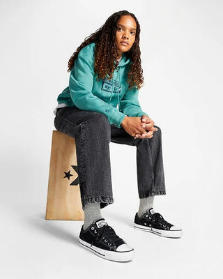 Person wearing a teal hoodie, gray jeans, and black sneakers sitting on a wooden stool.