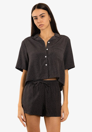 A woman in a Duvin Black Leopard Button-up Shirt with a relaxed, cropped cabana fit, made from a blend of 58% rayon and 42% polyester, featuring a camp collar, black leopard print, and side hem woven label.