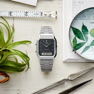 Silver Casio watch on a light wooden surface with a plant and measuring tape.