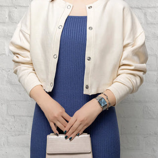 Person wearing a white cardigan over a blue dress, holding a beige handbag against a gray brick wall.