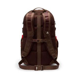 Close-up of the brown Nike ACG DAYMAX Backpack showing mesh paneling, adjustable webbing straps, buckle closures, and a red zipper pull