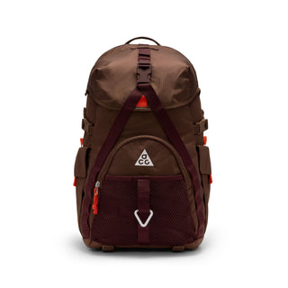 Close-up of the brown Nike ACG DAYMAX Backpack showing mesh paneling, adjustable webbing straps, buckle closures, and a red zipper pull