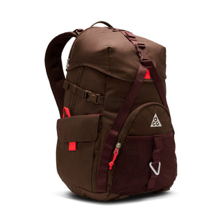 Close-up of the brown Nike ACG DAYMAX Backpack showing mesh paneling, adjustable webbing straps, buckle closures, and a red zipper pull