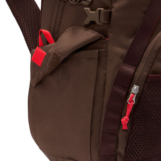Close-up of the brown Nike ACG DAYMAX Backpack showing mesh paneling, adjustable webbing straps, buckle closures, and a red zipper pull