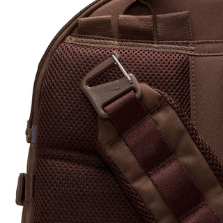 Close-up of the brown Nike ACG DAYMAX Backpack showing mesh paneling, adjustable webbing straps, buckle closures, and a red zipper pull