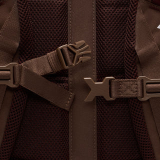 Close-up of the brown Nike ACG DAYMAX Backpack showing mesh paneling, adjustable webbing straps, buckle closures, and a red zipper pull