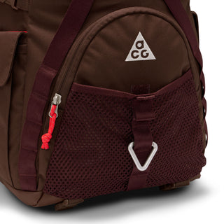Close-up of the brown Nike ACG DAYMAX Backpack showing mesh paneling, adjustable webbing straps, buckle closures, and a red zipper pull