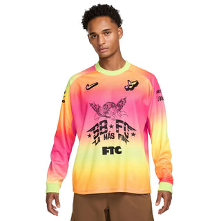 A male model wearing a long-sleeve Nike soccer jersey with a vibrant pink, orange, and yellow gradient design, featuring black graphics and logos including the Nike swoosh and 'SB FC' text.