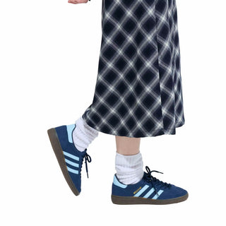 Person wearing blue Adidas sneakers with white socks and a plaid skirt on a white background