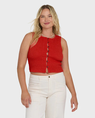 Woman wearing a red sleeveless top and white pants on a plain background