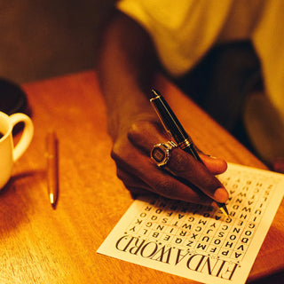 Person solving a crossword puzzle with a pen on a wooden table.