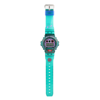 “Jae Tips G-SHOCK watch with translucent teal case and strap, lavender face, multicolor dial accents, flower graphics, and a red front button.”