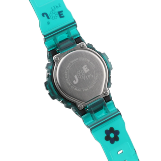 “Jae Tips G-SHOCK watch with translucent teal case and strap, lavender face, multicolor dial accents, flower graphics, and a red front button.”