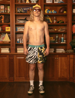 Man wearing colorful shorts standing in front of a bookshelf filled with various items.