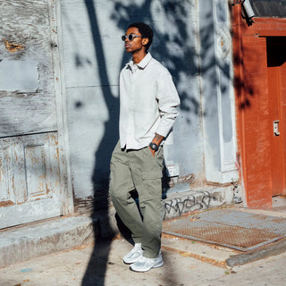 Man in light-colored shirt and green pants standing against a textured wall.