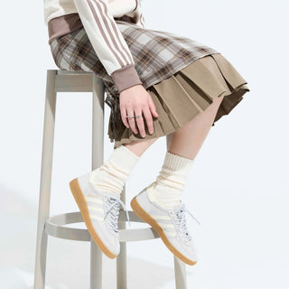 Person wearing a plaid skirt, white sneakers with brown soles, and beige socks sitting on a stool.