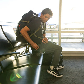 Model wearing Roark Layover Pro Pants with a backpack while seated in an airport terminal.