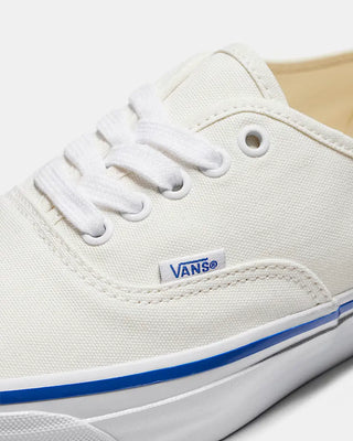 White Vans Authentic shoes with blue foxing stripe, white laces, and canvas uppers on a light neutral background.
