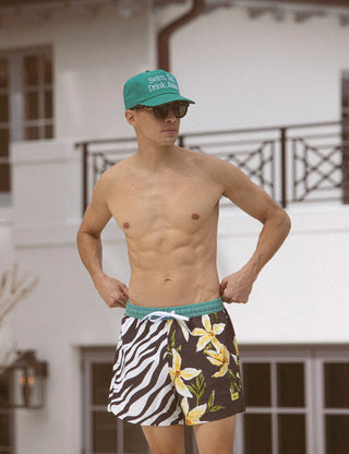 Man wearing a green cap and floral swim shorts standing in front of a building.