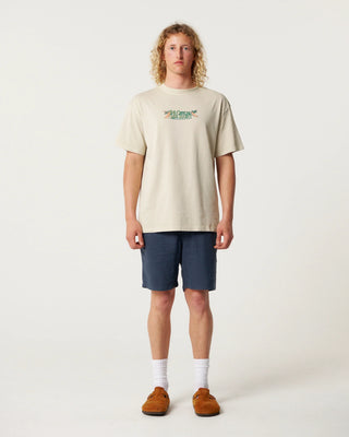 Person wearing a beige t-shirt with a logo and dark shorts on a white background