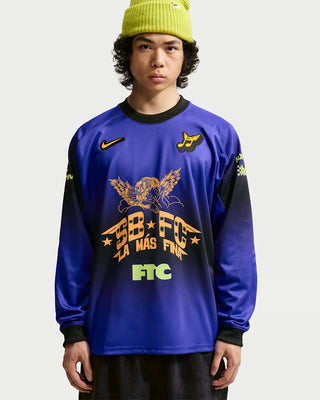 A model wearing a royal blue long-sleeve soccer jersey with black and yellow graphics, paired with a lime green beanie and black pants, against a light gray background.