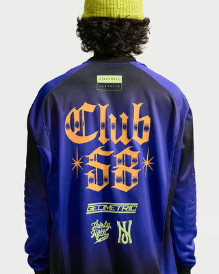 Back view of a person wearing a purple and black long-sleeve jacket with a large orange 'Club 56' graphic and smaller green and white graphics on the back, paired with a yellow beanie against a light gray background.