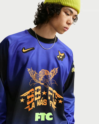 A close-up of a person wearing a royal blue long-sleeve soccer jersey with black sleeves, featuring a gold Nike swoosh, a winged eagle graphic, and 'SB FC LA MÁS FIT' text on the front, paired with a yellow-green beanie against a light gray background.