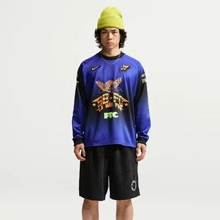 A model wearing a blue and black gradient long-sleeve athletic jersey with a large orange and yellow graphic on the front, paired with black shorts and a lime green beanie, against a plain light gray background.