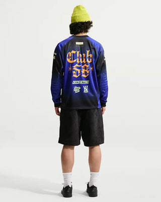 A person shown from the back wearing a blue and black long-sleeve jersey with a large orange and yellow 'Club 58' graphic on the back, paired with black shorts, white socks, and black shoes, against a light gray studio background.