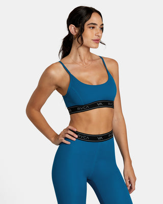 A confident woman wearing the RVCA Base Sports Bra, showing off the racerback design and half-moon back cut-out. The sports bra is made from recycled performance fabric.