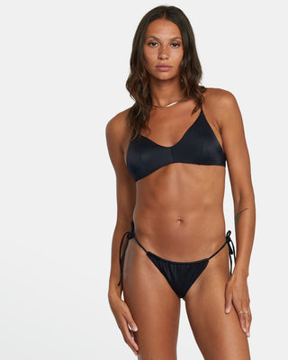 Stylish woman wearing black RVCA Solid Tie Skimpy Bikini Bottoms with adjustable side ties and cheeky cut, standing by the beach.