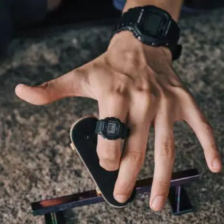 Black G-SHOCK DWN5600-1 ring watch with digital display on finger.