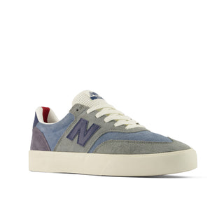 A New Balance low-top sneaker in a muted blue-gray suede and canvas mix, featuring a large navy 'N' logo on the side, white laces, and a thick cream-colored rubber sole.