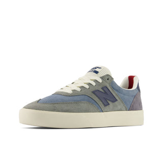 A New Balance sneaker in a muted colorway featuring a gray suede toe box, dusty blue suede overlays, a navy blue 'N' logo, off-white laces and midsole, with a small red accent visible inside the heel collar.