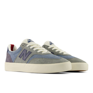 A pair of New Balance low-top sneakers in a muted blue-gray suede and canvas mix, featuring off-white laces and a thick off-white rubber sole, with a small red accent visible inside the heel collar.