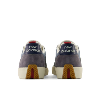 A pair of New Balance low-top sneakers shown from the back, featuring a navy suede heel tab with white 'new balance' branding, a dark gray suede upper, cream midsoles, and brown and red outsole patterns, set against a plain white background.