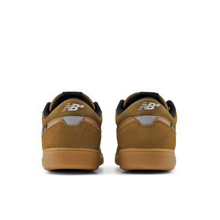A pair of New Balance sneakers shown from the back, featuring a mustard brown suede upper, black laces and collar, a white embroidered 'NB' logo, and a gum brown sole, set against a plain white background.