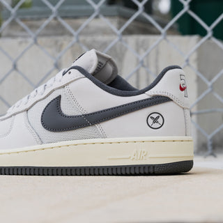 A Nike Air Force 1 Low sneaker in white and dark gray, featuring a cream midsole and a small red Nike logo on the heel, photographed outdoors in front of a chain-link fence.