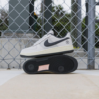 A Nike Air Force 1 Low sneaker in off-white and black, shown with its black outsole resting underneath the upper shoe, set against a chain-link fence background.
