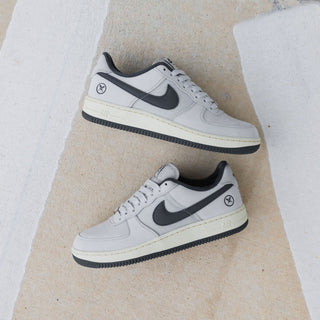 A pair of Nike Air Force 1 Low sneakers in light gray and black, styled on a textured beige concrete surface with white painted lines and a chain-link fence in the background.