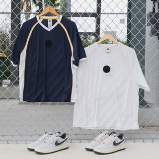 Two short-sleeve athletic t-shirts hanging on wooden hangers in front of a chain-link fence, with matching sneakers placed below them. The left shirt is navy blue with yellow shoulder stripes and a black circular chest logo.