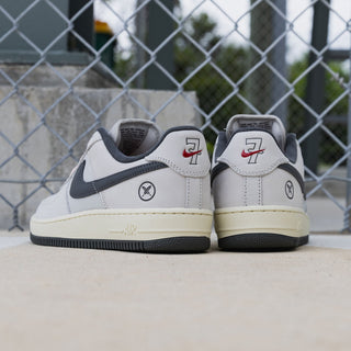 A pair of Nike Air Force 1 Low sneakers shown from the back, positioned on a concrete surface in front of a chain-link fence.