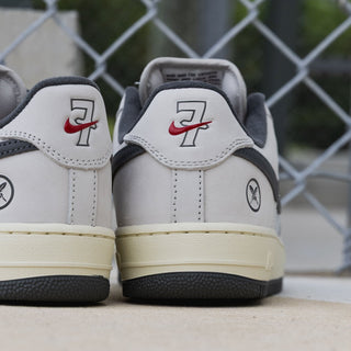A pair of low-top sneakers shown from the back, featuring a light gray leather upper with dark gray overlays, a cream midsole, and a black outsole. The heel tab displays a red Nike Swoosh and a black '7' logo, with a circular 'X' logo on the side heel.