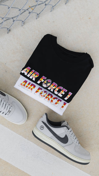A folded black T-shirt with a white hem and a colorful 'AIR FORCE' graphic, styled on a light beige surface with two gray sneakers nearby.